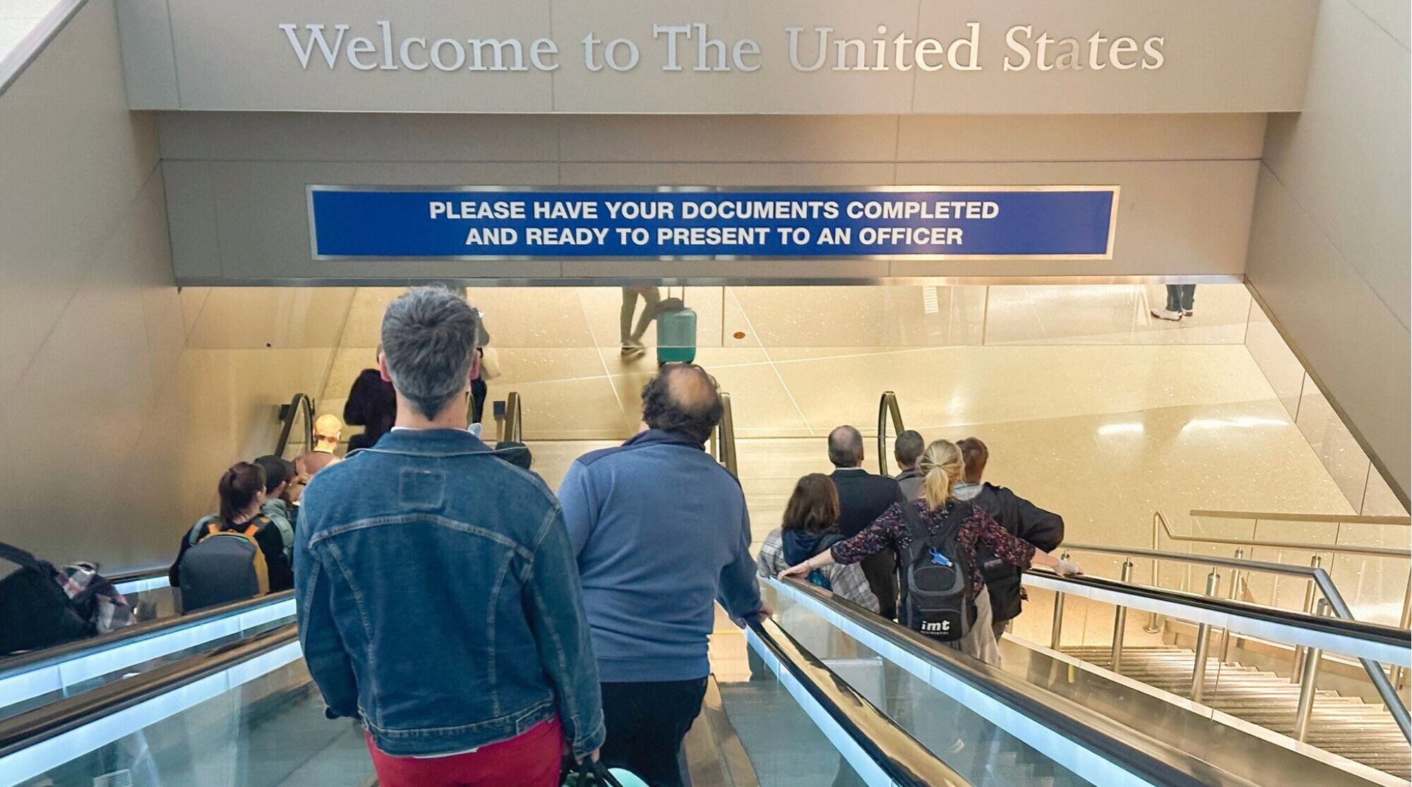Passageiros caminhando em direção à Alfândega e Proteção de Fronteiras dos EUA no Aeroporto Internacional de Los Angeles com uma placa de 'Bem-vindo aos Estados Unidos'