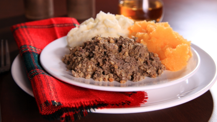 A close-up coloured image of a white plate with a red piece of Scottish tartan next to it, to be used by a diner as a napkin. On the plate are three small piles of different foods - neeps and tatties and a pile of minced haggis. To illustrate a blog post entitled 'What is Burns Night and How to Celebrate It'.