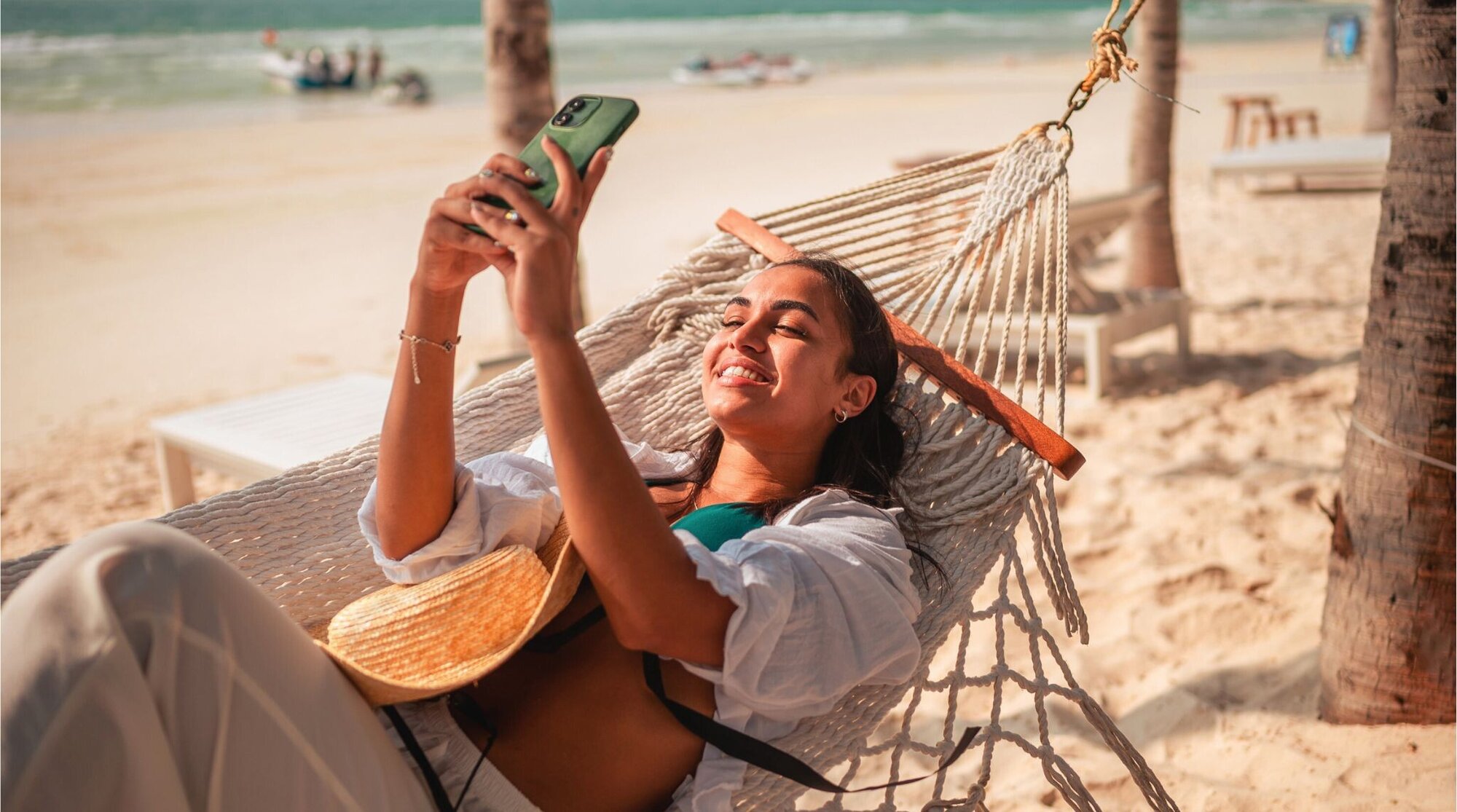 Mulher usando celular enquanto descansa em uma rede na praia, com o mar ao fundo.