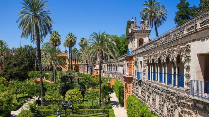 Seville’s Real Alcázar gardens in Spain, filled with tall palm trees, carved stone arches, and flower-filled courtyards under a clear blue sky. A cultural city break option for where is hot in October in Europe, combining history, warmth, and the best eSIMs for travel.