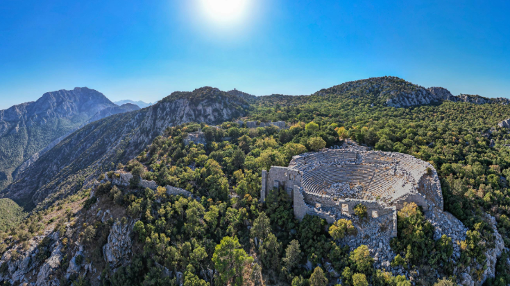 A photograph of Turkey's Taurus mountain range covered in lush greenery against a blue sky. In the foreground is the Termessos ancient city, showing the amphitheatre. Termessos is one of Antalya and Turkey's most outstanding archaeological sites. To illustrate a blog post entitled 'nine things to do in Antalya'.