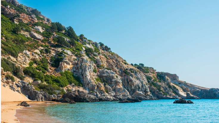 Wide sandy beach with gentle waves lapping at the shore in Crete, Greece. Behind the beach rise rugged golden cliffs dotted with green shrubs — a warm escape for anyone asking where is hot in October in Europe, ideal for cheap hot holidays in October with travel eSIMs.