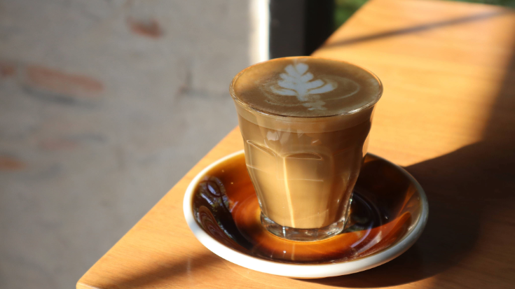 A photograph of a small glass on a saucer, sitting on a table by a window. The sun is streaming through the window and onto the glass, which contains a flat white coffee. On the surface of the medium-brown-coloured coffee is some white latte art in the form of a decorative leaf. To illustrate a blog post entitled 'Which Country Has the Best Coffee Culture?'