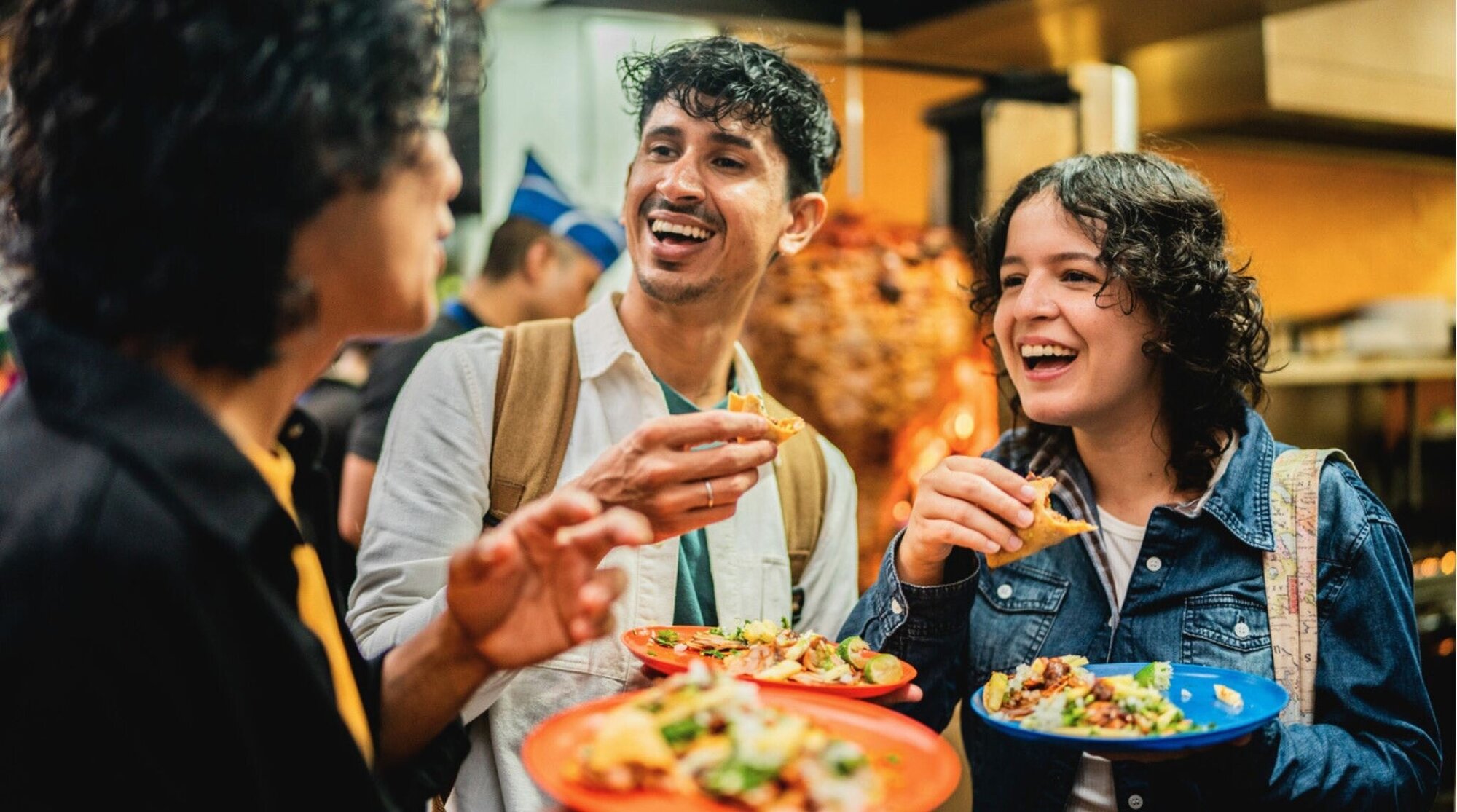 Grupo de amigos riendo y comiendo tacos al aire libre, ambiente de viaje y comida callejera.