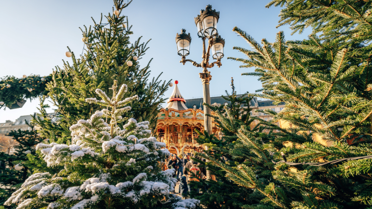 A colour image showing a row of Christmas trees dusted with snow and an old Victorian lamp post in the foreground. In the background is a blue daytime crisp winter sky and a traditional gold and red merry-go-round fairground ride in front of Hotel de Ville, the city hall of Paris. To illustrate a blog post entitled 'Extreme Day Trips: Eurostar Christmas Markets Edition'.