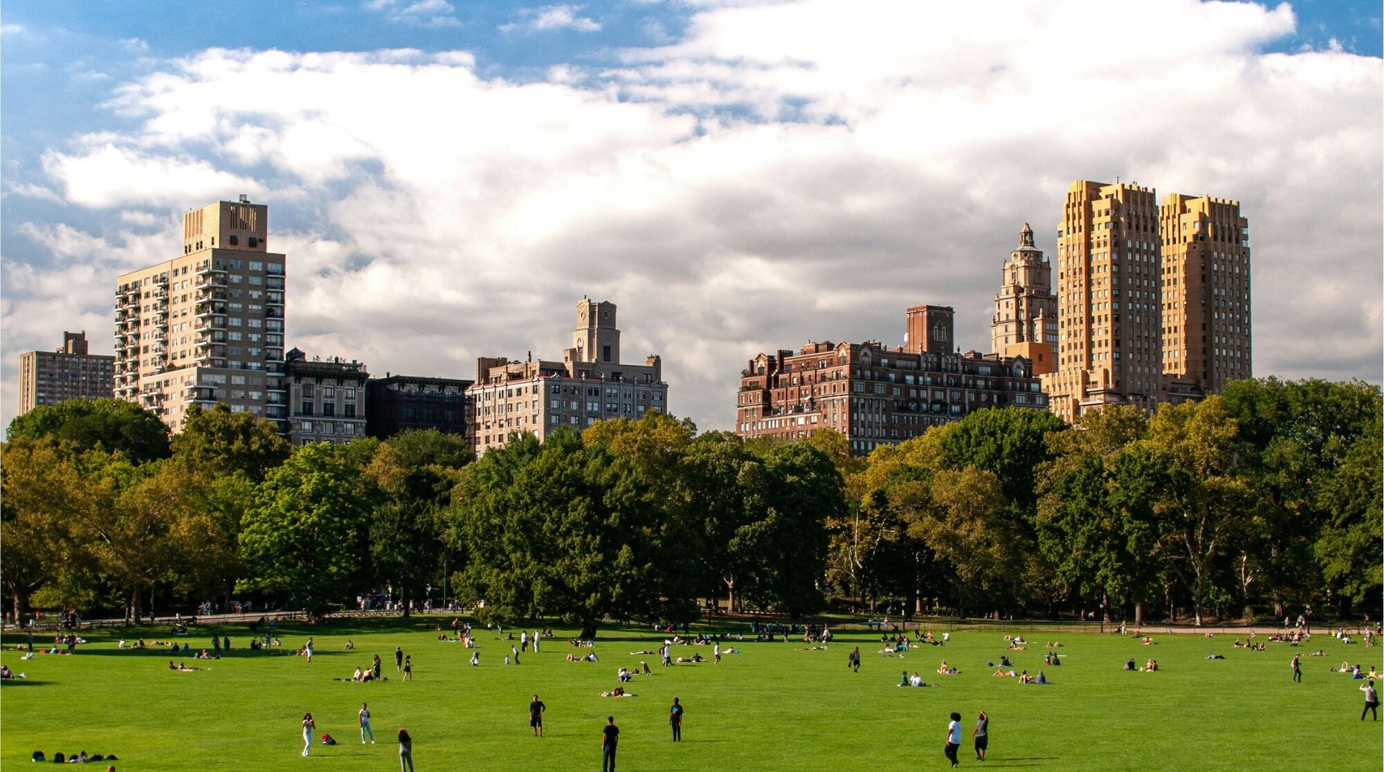 Vista de turistas e nova-iorquinos relaxando ou brincando no gramado do Central Park, e em segundo plano, prédios da cidade no horizonte.