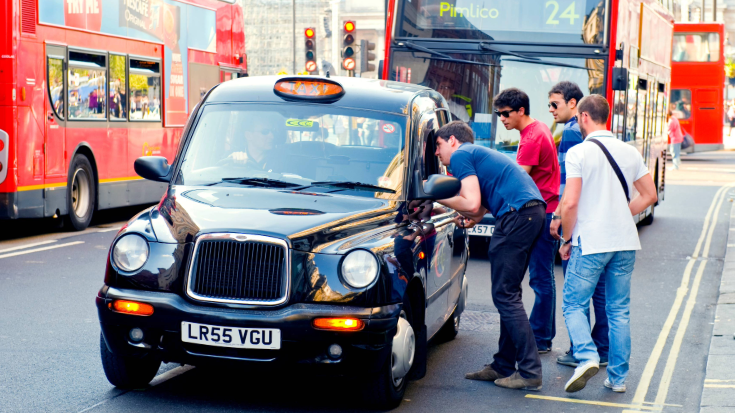 Four young men in t-shirts speak to the driver of a London black cab, with a bright red London bus in the background.