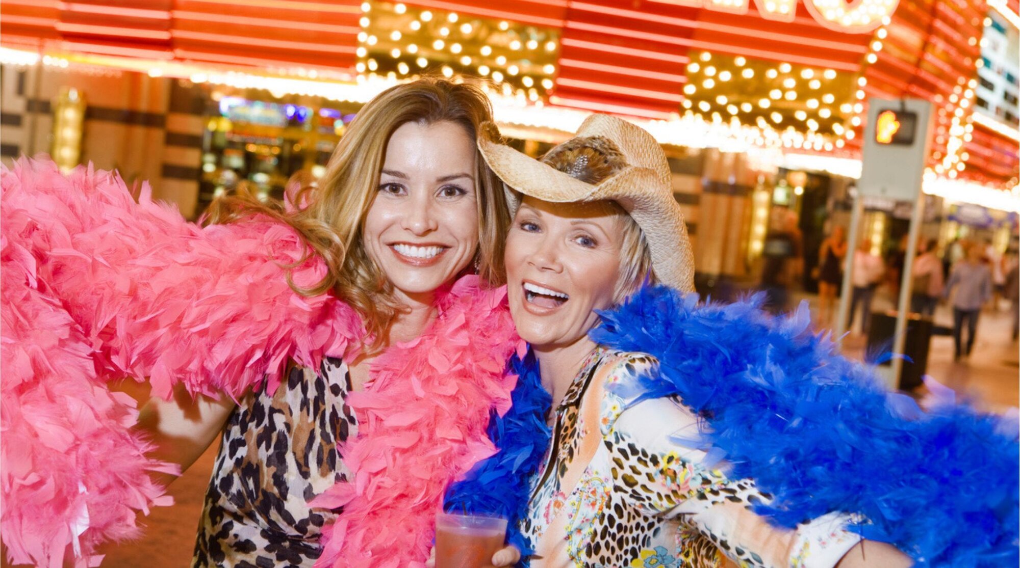 Dos mujeres posando sonrientes de noche con plumas de colores, con luces de casino y letreros al fondo.