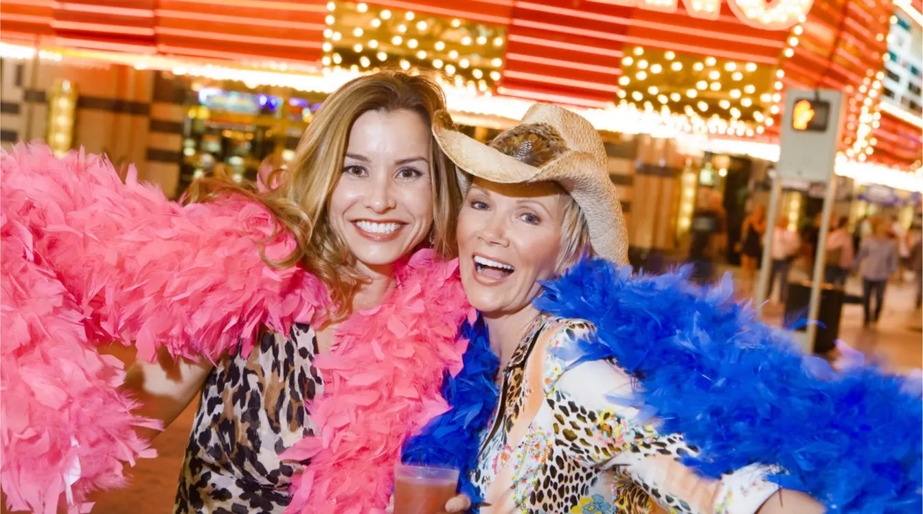 Dos mujeres posando sonrientes de noche con plumas de colores, con luces de casino y letreros al fondo.