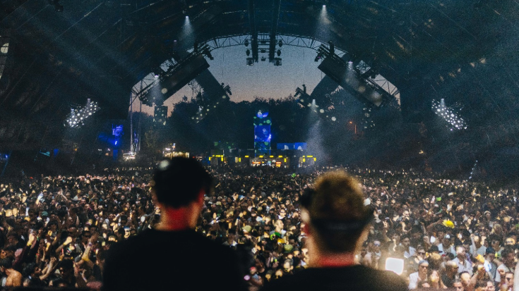 A photograph showing the blurred backs of two people's heads as they stand and perform on a large stage in the foreground. In the background, a large crowd has gathered and is listening to the music. To illustrate a blog post entitled 'The Ultimate Sziget Festival Guide.'
