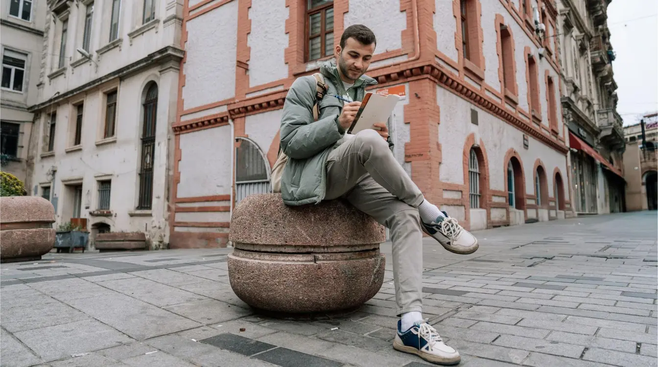  Um jovem viajante, de jaqueta verde e mochila, sentado em um bloco de pedra, em uma rua de paralelepípedos, em Istambul, Turquia. Ele segura uma caneta e escreve em um caderno, refletindo sobre sua viagem cercado por prédios históricos.