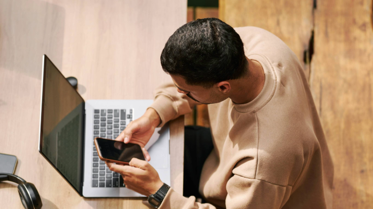 A photograph os a man from above. He is sitting at a wooden desk or table in front of an open laptop. He is looking at his mobile phone and appears to be thinking or doing research. He is wearing a beige jumper and has black hair and a black moustache. This is an image to illustrate a blog post entitled 'Unlimited eSIM: Plans, Benefits, and How It Works'.