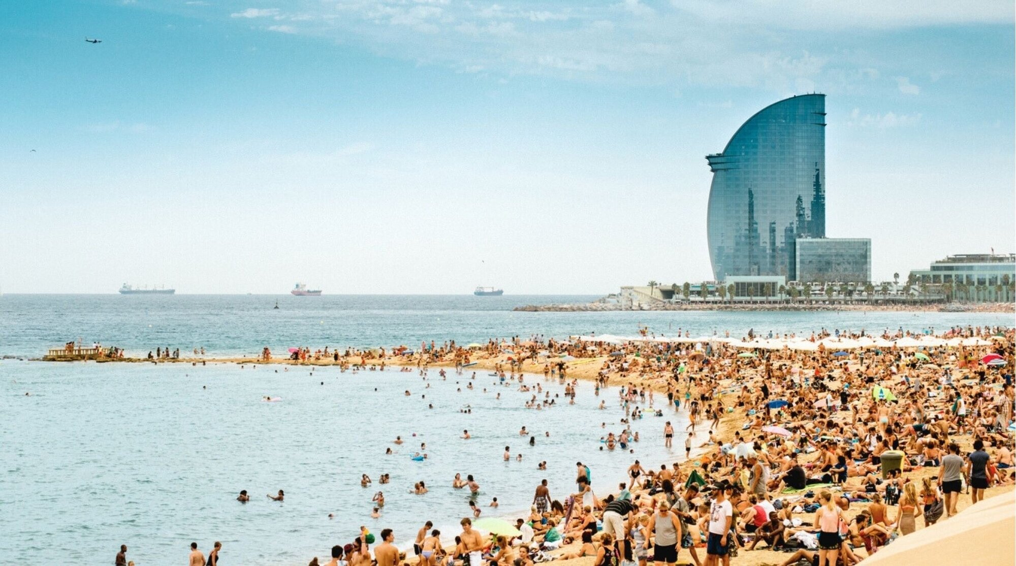 Playa de la Barceloneta llena de gente con el icónico hotel vela al fondo en Barcelona