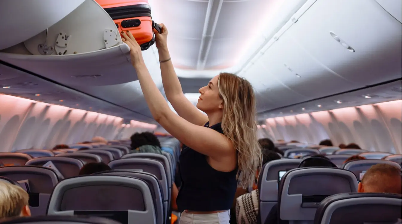 Mulher de cabelos claros dentro da cabine de um avião, guardando uma mala de mão de cor laranja no compartimento de bagagem superior.