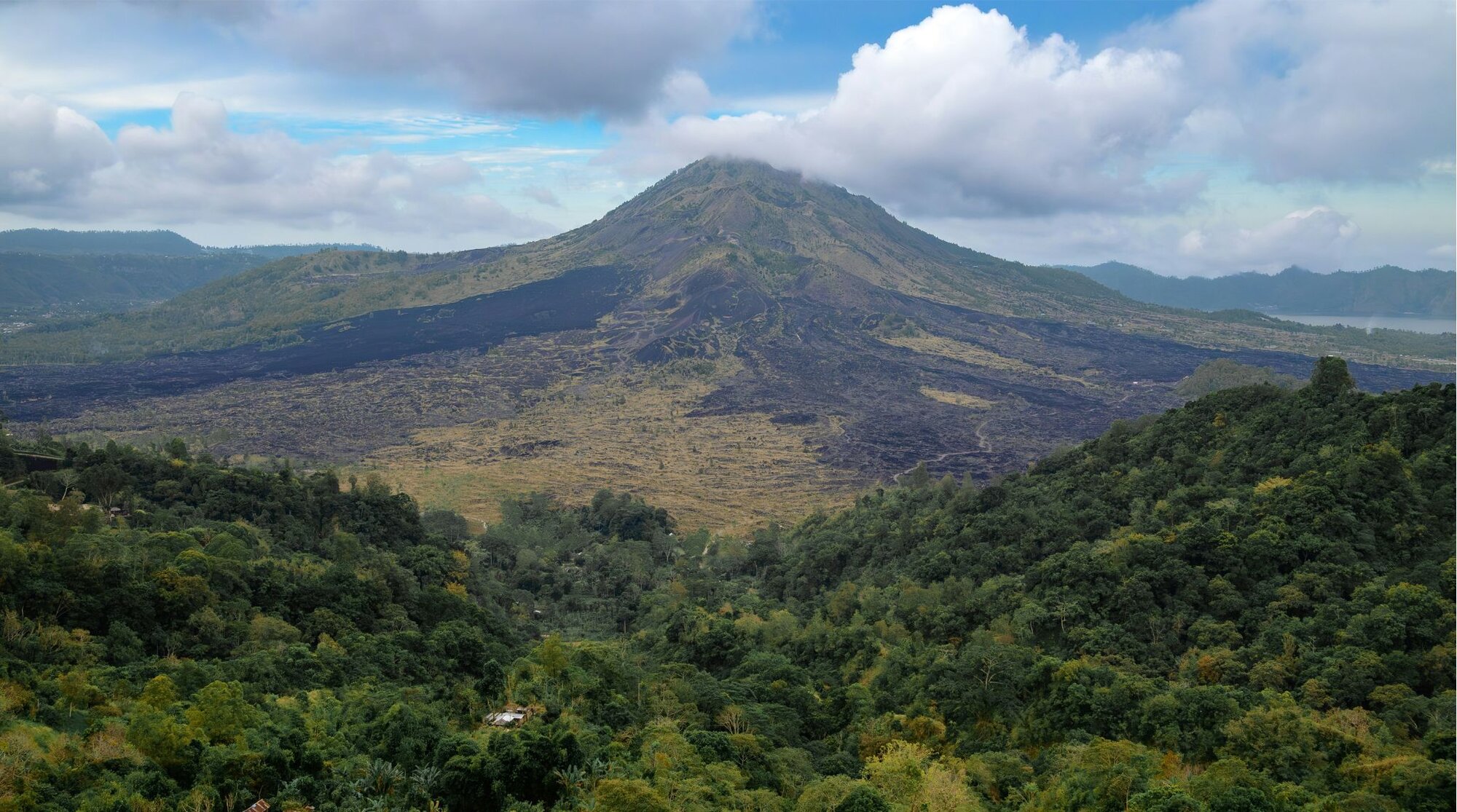 View of Mount Batur from a cafe in Kintamani, overlooking a lush forest and the black slopes of the volcano.