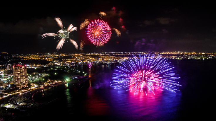 An image of a coastal seaside town in Okinawa, Japan, at night. Three very large yellow, white and blue fireworks are exploding against the pitch black sky. To illustrate a blog post entitled 'Top Things to Do in Okinawa, Japan's Tropical Gem.' 