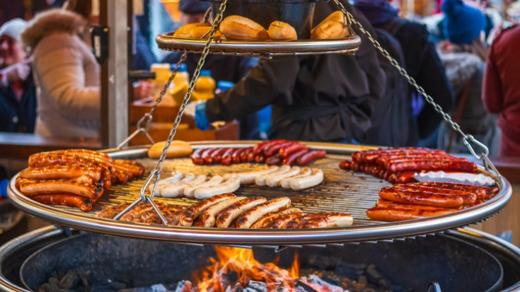 A photograph of a German-style, hanging circular grill full of bratwurst sausages grilling away. Some are red, some are golden brown. To illustrate the Winter Wonderland Christmas Market at Hyde Park in a blog post entitled 'Things to do in London in December'.