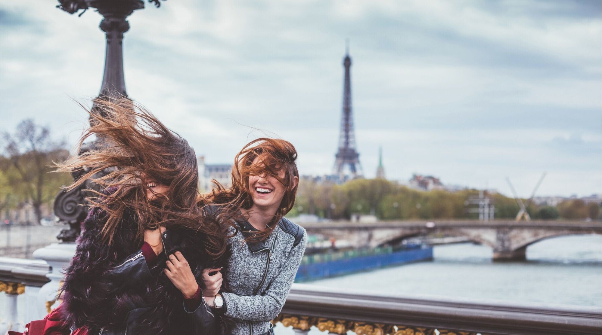Duas jovens amigas caminhando por Paris enquanto o vento levanta seus cabelos, com a Torre Eiffel ao fundo.