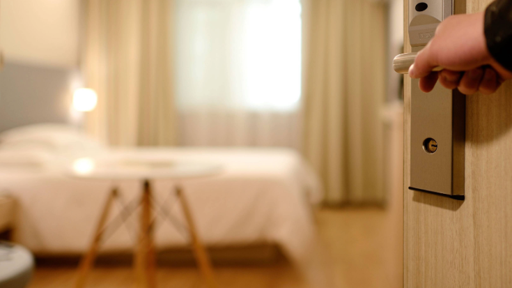 A photograph of an image of a blurred-out and clean, softly-lit hotel room in the background and in the foreground, there is a hand opening a door handle, which leads into the room.
