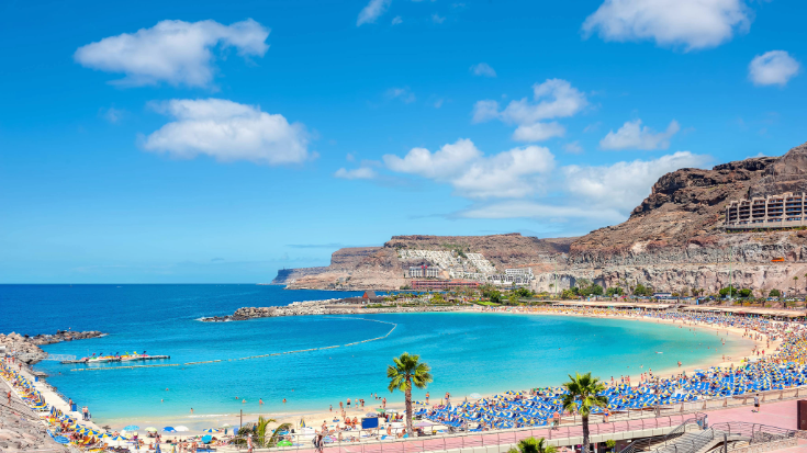 An image of a crowded pale sandy beach in Gran Canaria in Spain with bright blue skies and mountains in the background to illustrate a blog post entitled The Best Winter Family Holidays from Ireland. 