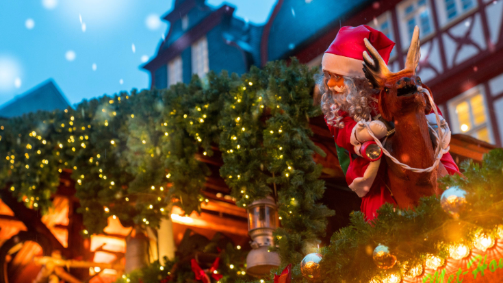 An image of a toy doll of Santa Claus sitting on the roof of an alpine hut at a Christmas market, illustrating a blog post entitled The Best Christmas markets in the UK for 2024.