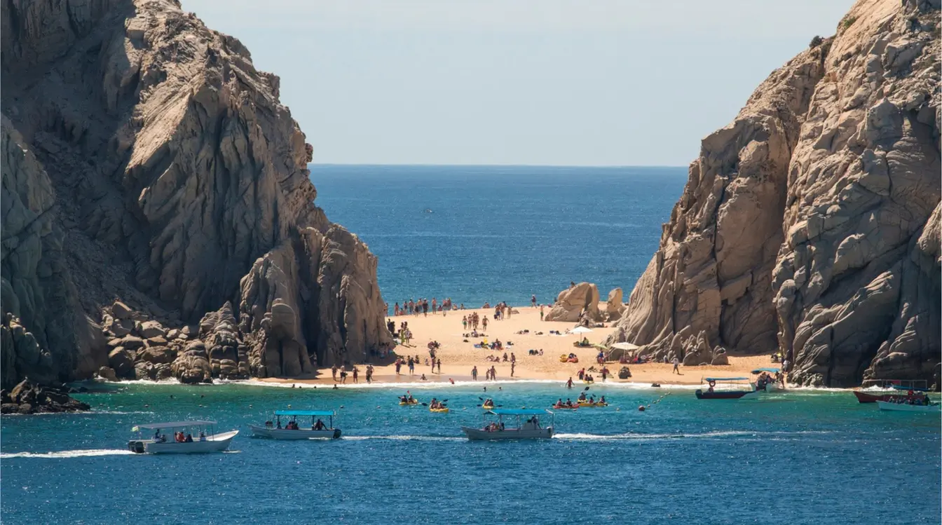 Vista del mar azul entre formaciones rocosas, con personas al fondo en una playa de Los Cabos.