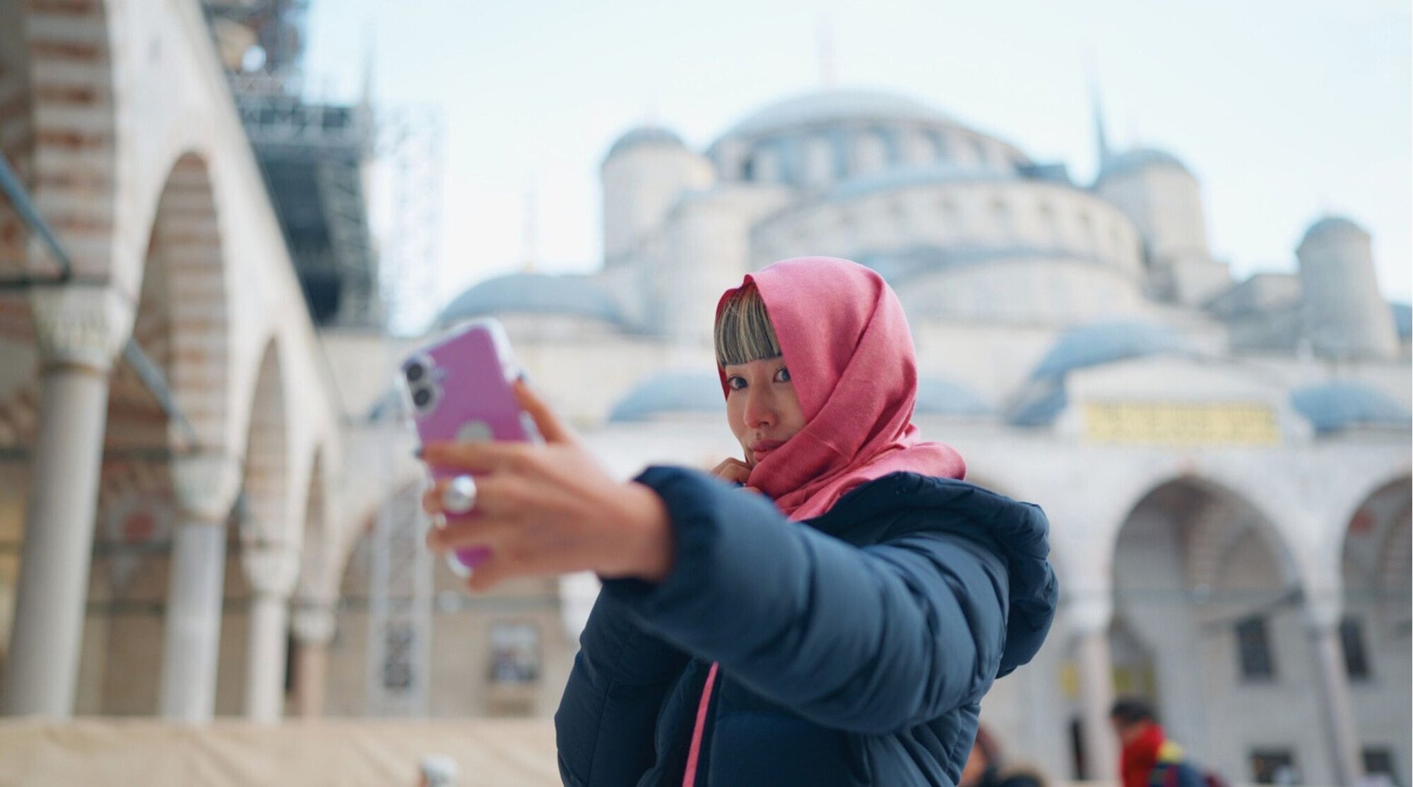 Uma viajante solo, usando um lenço rosa, tira selfies com seu smartphone tendo a Mesquita Azul ao fundo, no pátio frontal da mesquita em Istambul, Turquia.