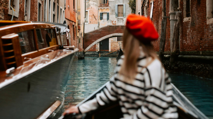 A photograph with a blurred-out image of a woman in the foreground turning and looking behind her. All the viewer can see of her is a red beret on her head, long brown hair and a white jumper with black horizontal stripes. In the background is a canal, probably in Venice, Italy and she is on a gondola. Either side of her are high walls in red brick in the romantic architectural style of Venice. This is an image to illustrate a blog post entitled 'Unlimited eSIM: Plans, Benefits, and How It Works'. 