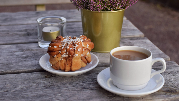 A photograph showing a wooden table outdoors. On the table is a vase filled with pink heather, a plain white coffee in a white cup and a Swedish cinnamon bun. The bun has flecks of brown cinnamon running through its plaits and also pearl sugar studding its surface. To illustrate a blog post entitled 'Which Country Has the Best Coffee Culture?'