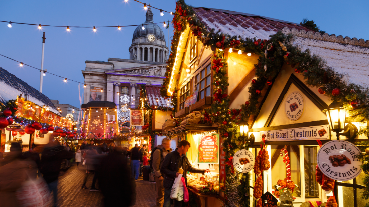 A colour photograph of a man making purchase at Nottingham Christmas Market. You can also see Christmas lights and small german-style chalet huts decorated with red and green garlands and golden bells. To illustrate a blog post entitled 'Extreme Day Trips - UK Christmas Markets Edition'.