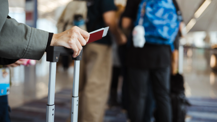 A photograph showing a close-up of a man's hand leaning on a suitcase handle, holding a British passport. He is waiting in a queue at the airport. To illustrate a blog post entitled ''Will ETIAS and EES Delay You? What UK Travellers Need to Know'?'