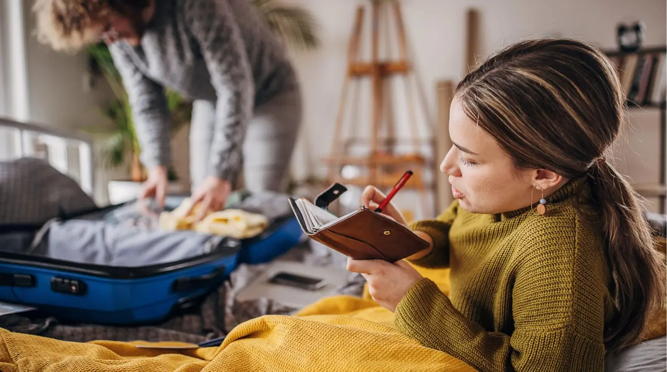 Casal se preparando para a viagem: mulher sentada na cama, anotando um checklist em um caderno, enquanto o homem arruma a mala azul ao fundo.