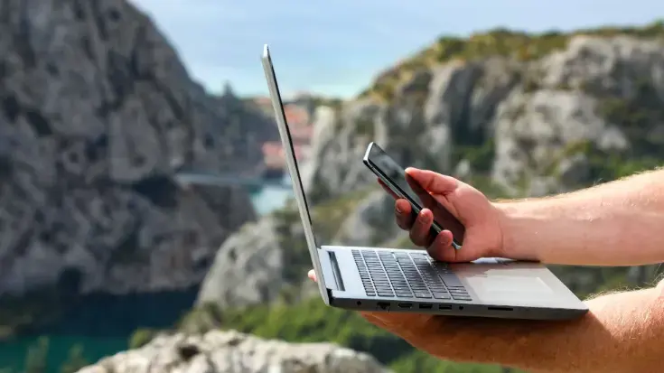 A close-up of a traveller's hands holding a laptop and smartphone against a scenic mountain backdrop, illustrating the importance of researching sustainable travel options online.