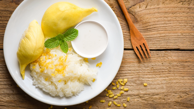 An image of some Thai sticky rice on a white plate, next to the bright yellow flesh of the durian fruit, topped with a mint leaf, to illustrate a blog post entitled '33 fun facts about Thailand' and the fact that this fruit is banned from many public places in Thailand, such as hotels and airplanes.