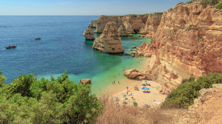 An image from a clifftop looking out over Praia da Marihna beach on the Algarve Coast in Portugal, the cliffs are a sandy orange colour, the sea and sky are bright blue, it's a sunny day and people on the beach below are relaxing and paddling in the water, to illustrate a blog post entitled' 'Where is hot in October in Europe?' A small Algarve cove in Portugal, where sunbathers relax on soft golden sand surrounded by towering limestone cliffs. The turquoise water is calm and clear, making this a classic choice for hot holidays in October and where is hot and cheap in October.