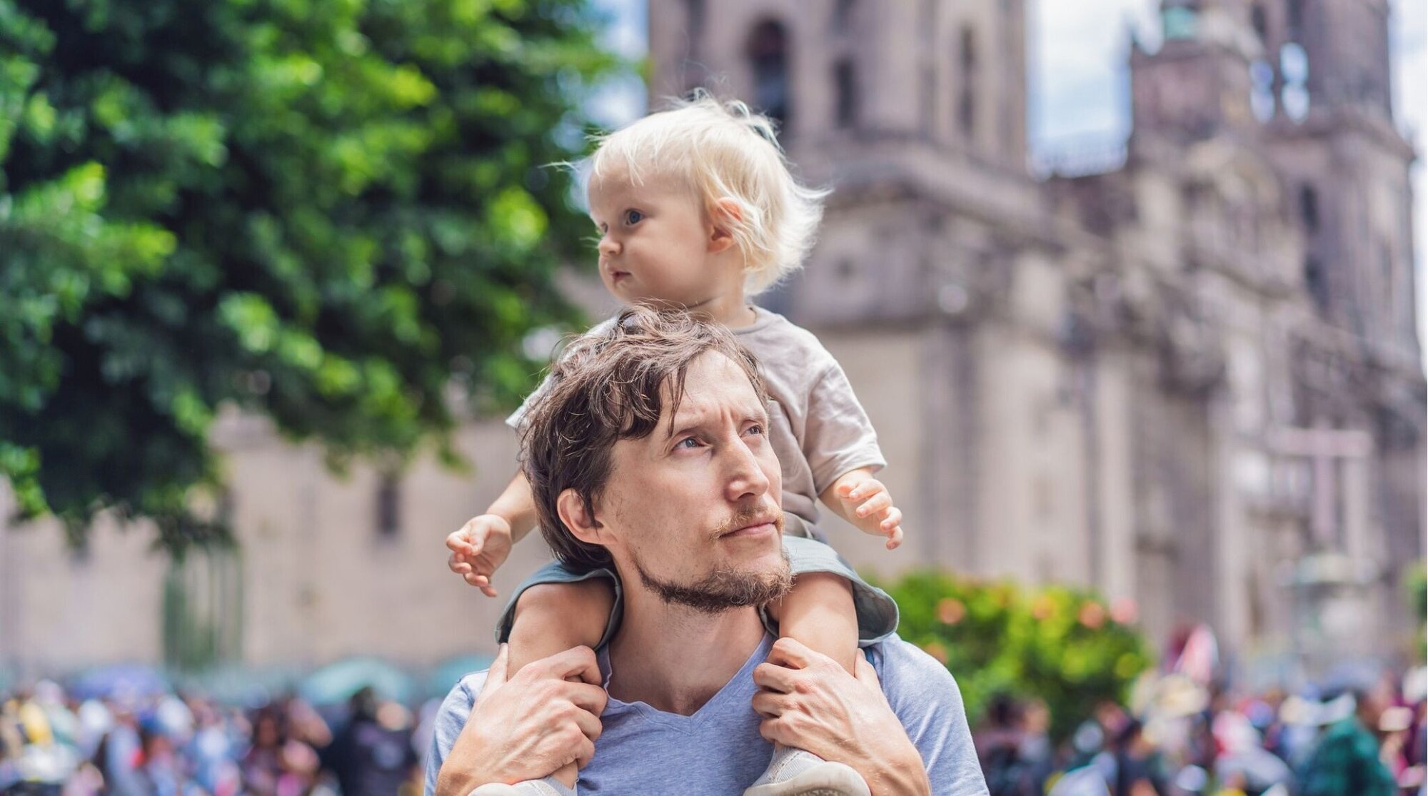 Un hombre con un niño sentado en los hombros observa una catedral al fondo, en una plaza concurrida.