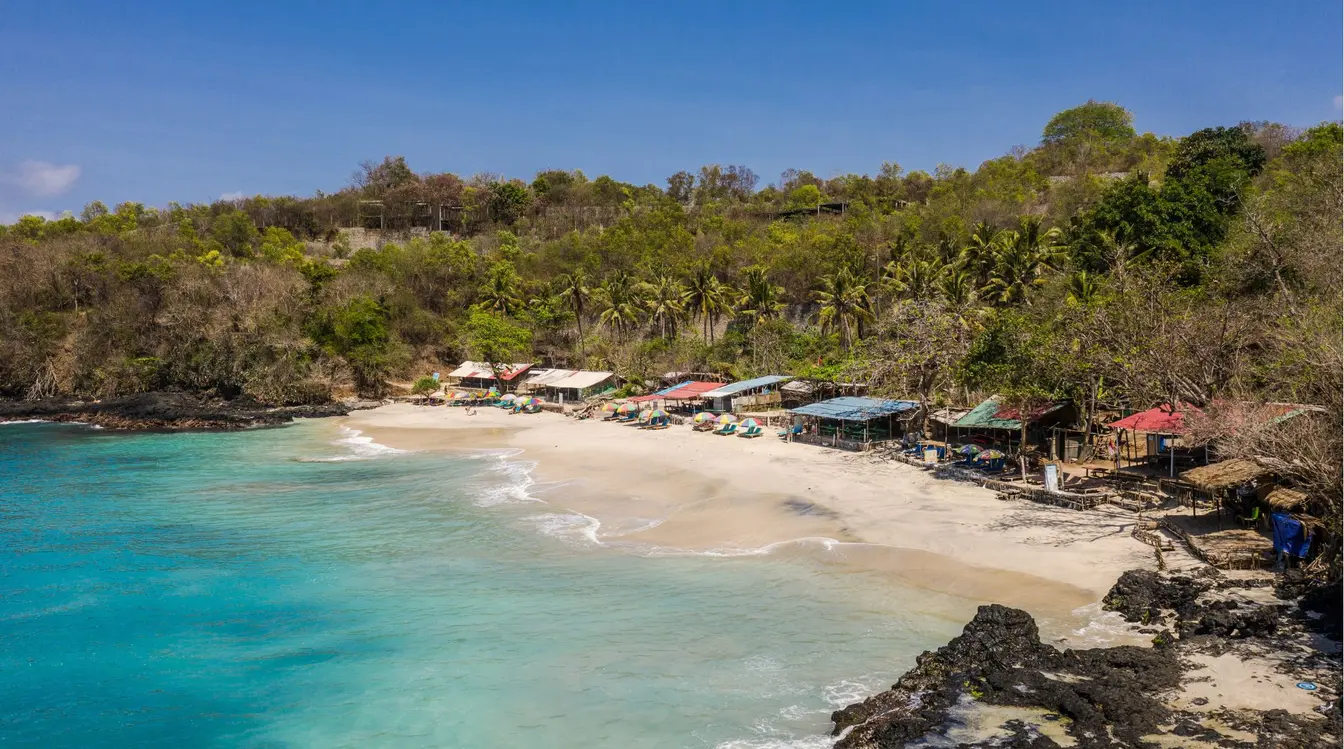 View of Bias Tugel Beach near Candidasa, with turquoise sea, a sandy shore, some beach shacks, and a few sunbeds.