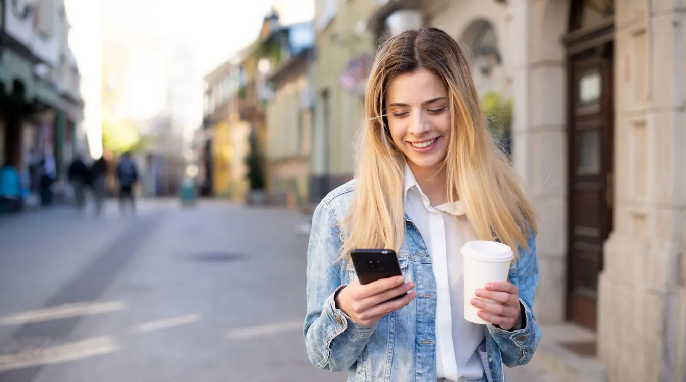 A UK woman in a blue jacket holding a phone, happy on holiday abroad after figuring out how to avoid roaming charges