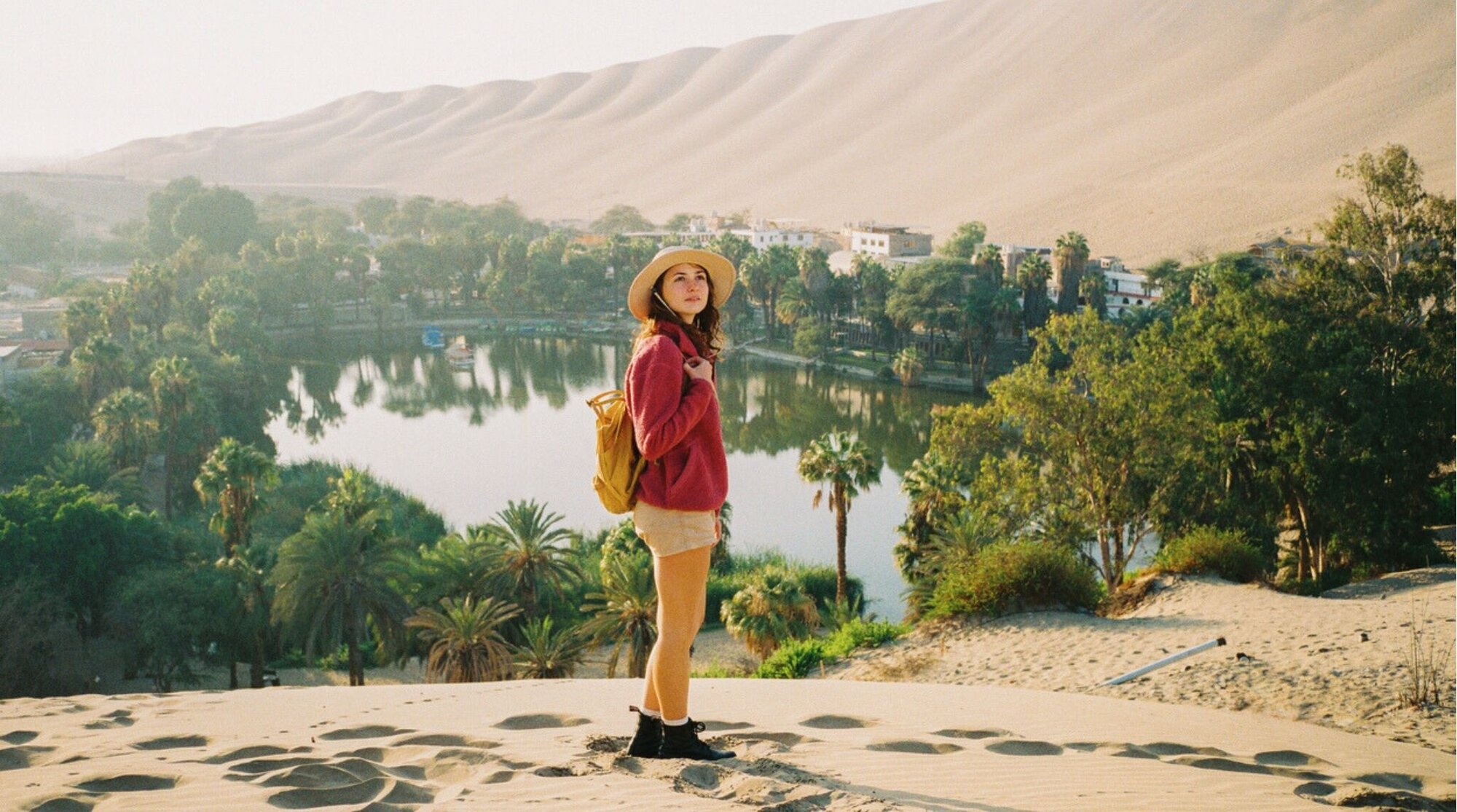 Viajera de pie sobre una duna con vistas a un oasis, paisaje desértico y palmeras al fondo.