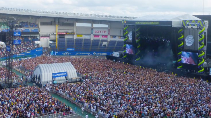 An aerial view of a large outdoor music festival grounds with a stage, showing the scale of the Summer Sonic Music Festival in Chiba and Osaka.