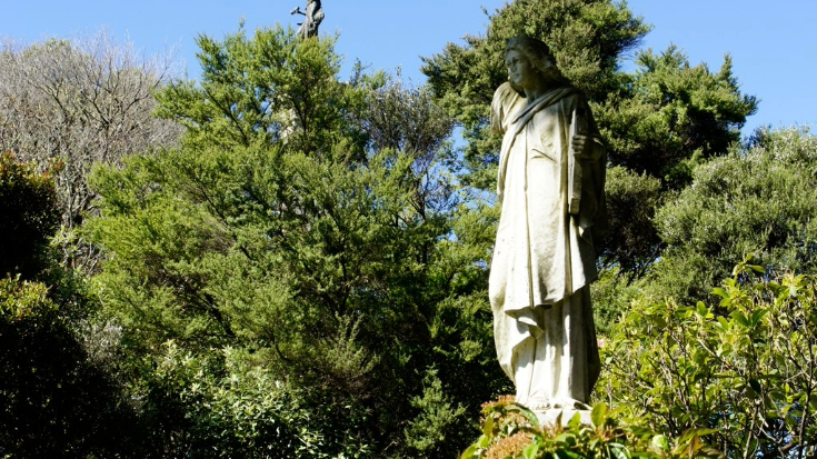 A colour photograph of Bolton Street Cemetery in Wellington, New Zealand. There are fir trees with full green foilage in the background and in the foreground is a sand-colourd statue of a woman, partially covered by the leaves. The sky is bright. blue, it's a sunny day and the cemetery appears pleasant and peaceful. To illustrate a blog post entitled '10 of The World's Most Beautiful Cemeteries'.
