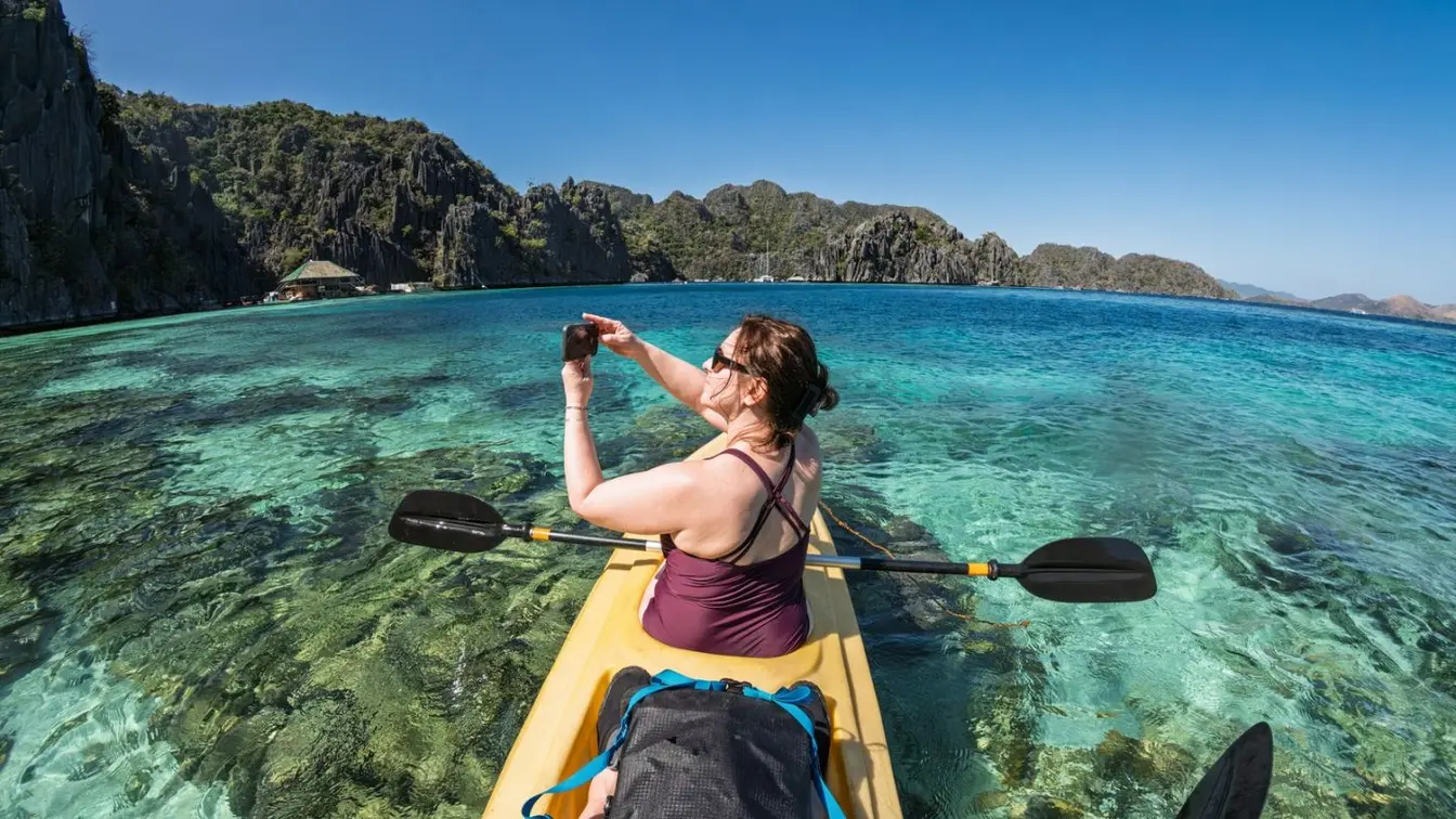 A woman taking a photo on her phone while kayaking through the turquoise sea in Coron, Palawan.
