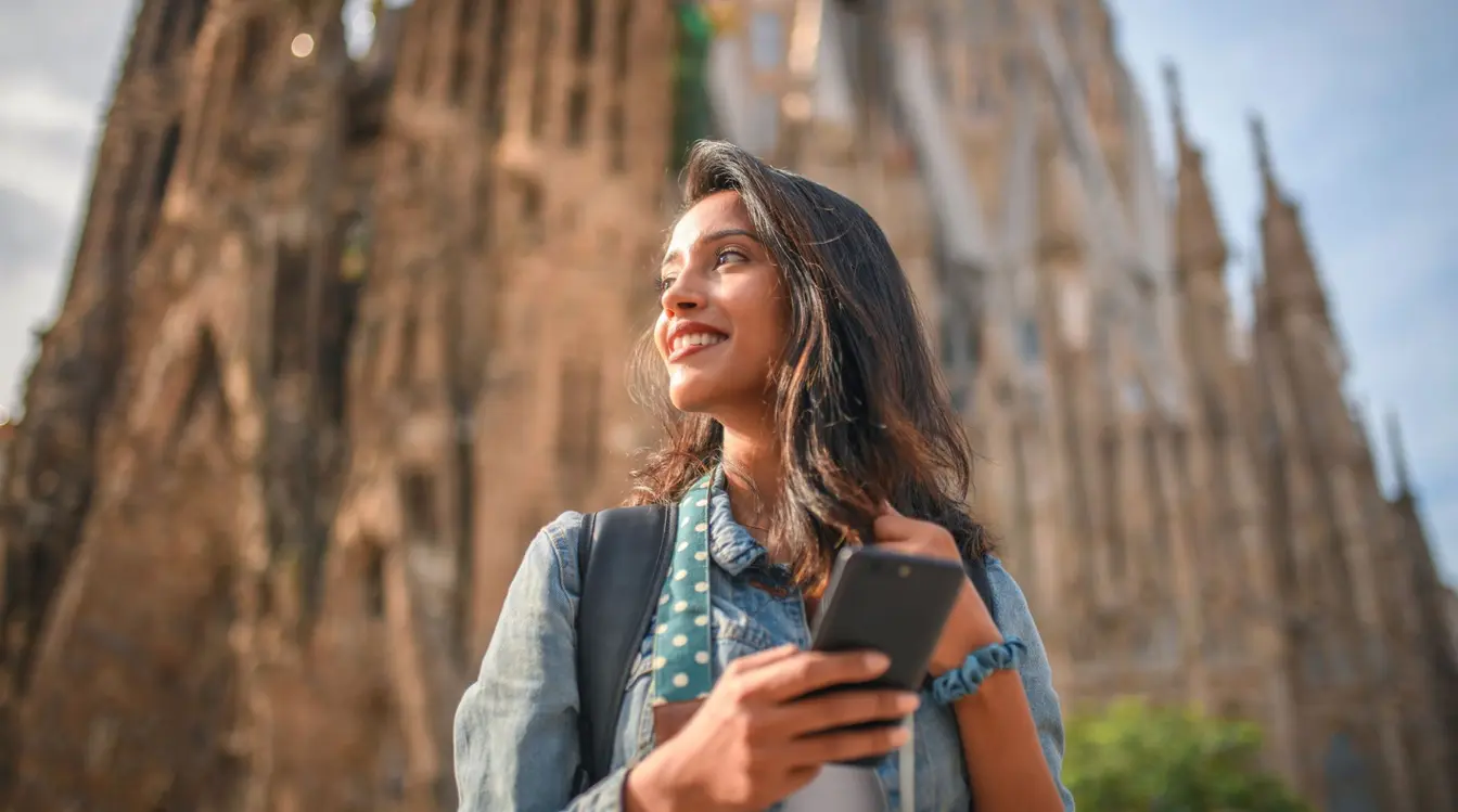A smiling woman holding a smartphone in front of the Sagrada Familia in Barcelona.