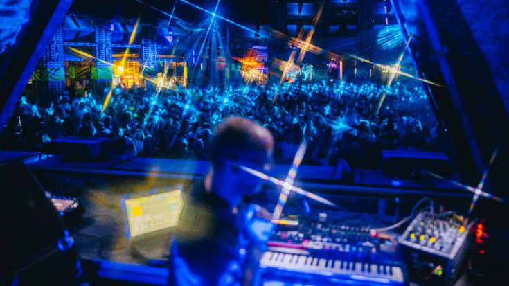 A photograph os a DJ on the stage in the foreground, he is blurred out slightly so you can't see his face but his right-hand profile is turned towards the camera. In the background is a concert crowd, slightly blurred also, with orange bokeh and blue lights illuminating the scene. To illustrate a blog post entitled 'The Ultimate Sziget Festival Guide.'