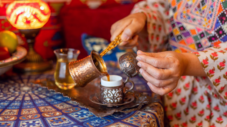 A photograph of a close up of a Turkish woman's hands One hand is pouring a thick brown coffee from a small copper pot with a long handle. The other hand is holding a traditional decorative copper Turish coffee cup, which is adorned with intricate patterns. In the background is a table with a tablecloth coloured in blues, reds, oranges and yellows. for a blog post entitled 'Which Country Has the Best Coffee Culture?'