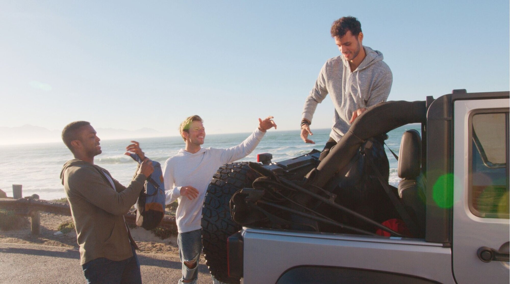 Tres amigos descargando equipaje de un Jeep descapotable en un viaje por carretera junto al mar.