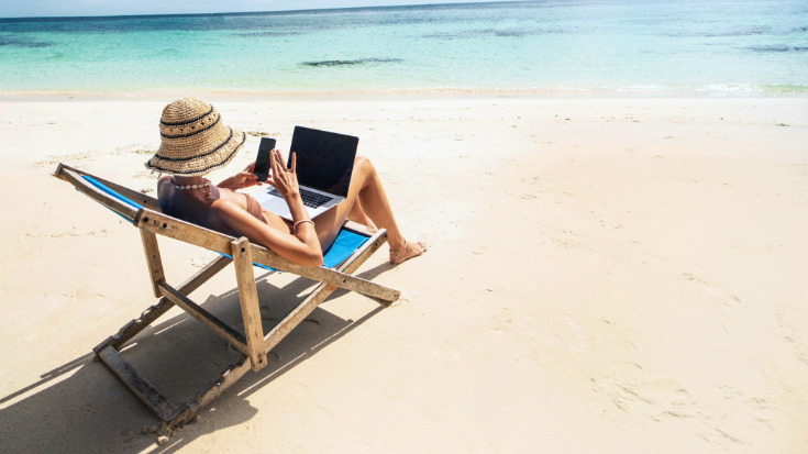 A woman in a sun hat photographed from the side, sitting in a deck chair and looking at her tablet, with her laptop balanced on her knees. She is sitting on a beautiful white sand beach and in the background is a calm sea with crystal waters and a blue cloudless sky. To illustrate a blog post entitled 'where is hot in October in Europe?'