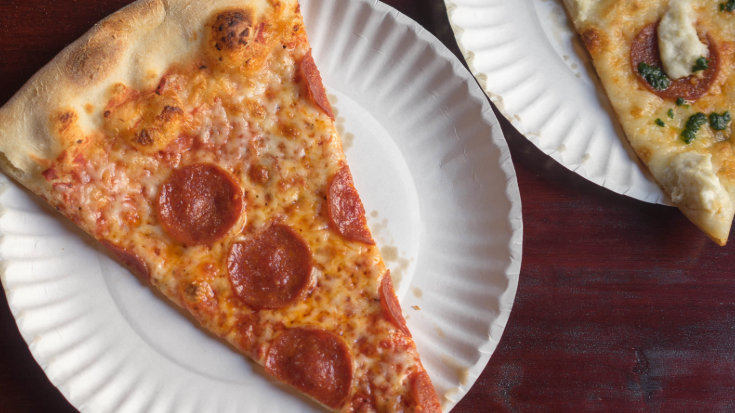 A close-up coloured photograph of a typical New York slice of pizza. It is large and is topped with cheese and pepperoni, to illustrate a blog post entitled 'New York Comic Con 2025: A Complete Travel Guide for Attendees'.