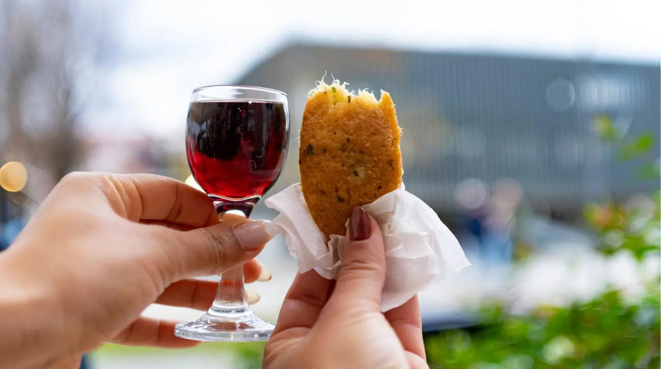 Duas mãos em primeiro plano contra um fundo urbano desfocado ao ar livre. A mão da esquerda segura uma pequena taça de vinho do porto. A mão da direita segura um bolinho de bacalhau frito dourado, parcialmente mordido.