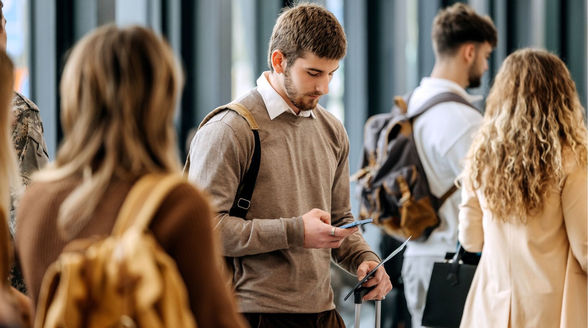 Viajante em fila no aeroporto segurando passaporte e celular, com outros passageiros ao fundo.